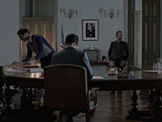 Image of three men in a room around a desk, a scene from the film 'The Secret Agent'