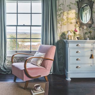 Accent chair next to a chest of drawers in a room with floral wallpaper and a view out into the countryside