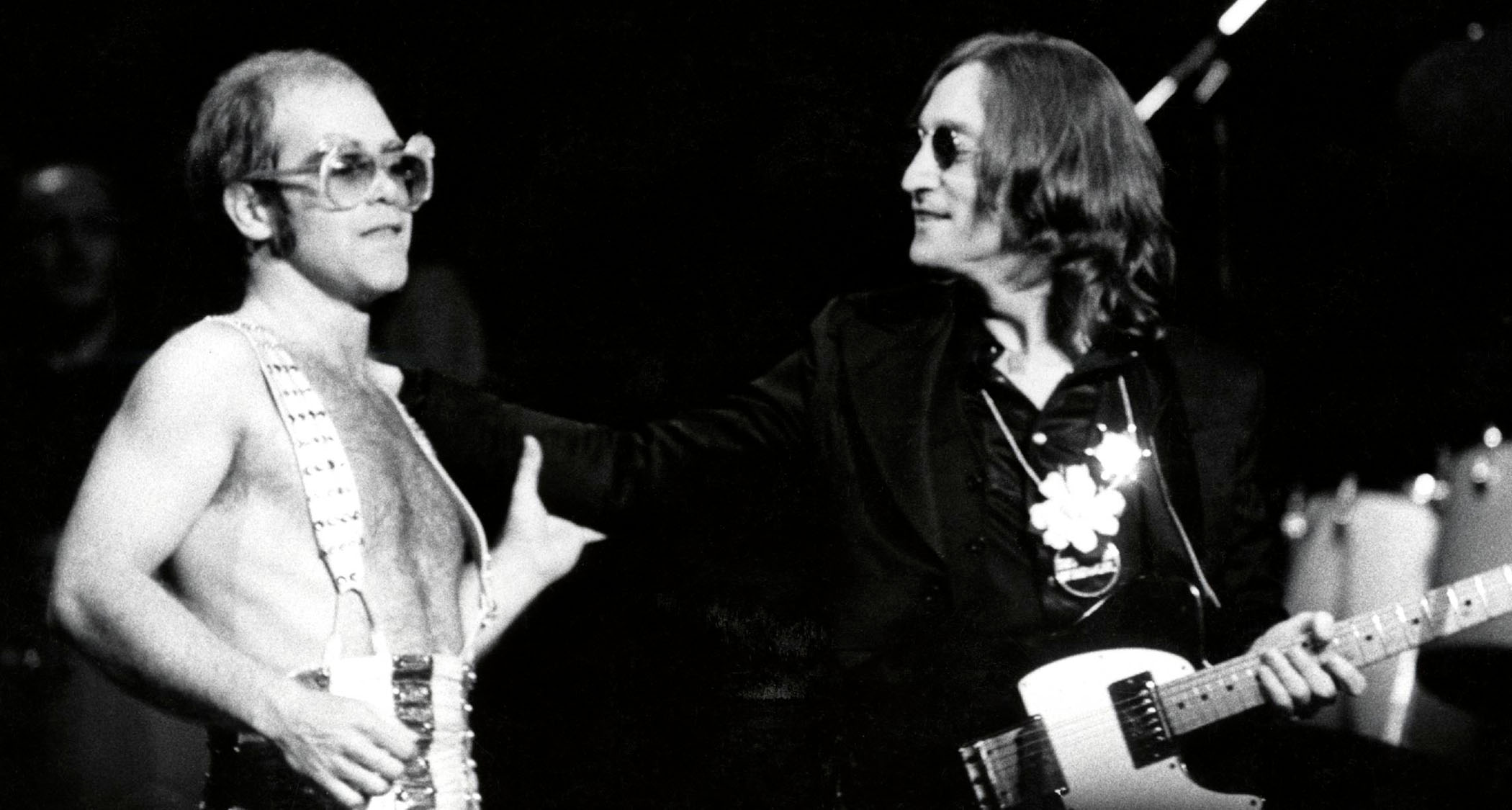 Elton John, bare chested but wearing braces and custom sunglasses, performs with John Lennon at his Madison Square Garden Thanksgiving show in 1974. Lennon plays a Fender Telecaster Deluxe.
