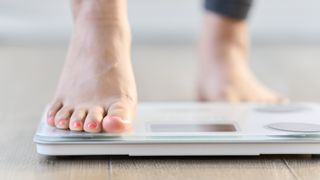 Close-up of someone placing one of their feet on a weighing scales. The foot on the scales is in focus, while the background is blurred. 