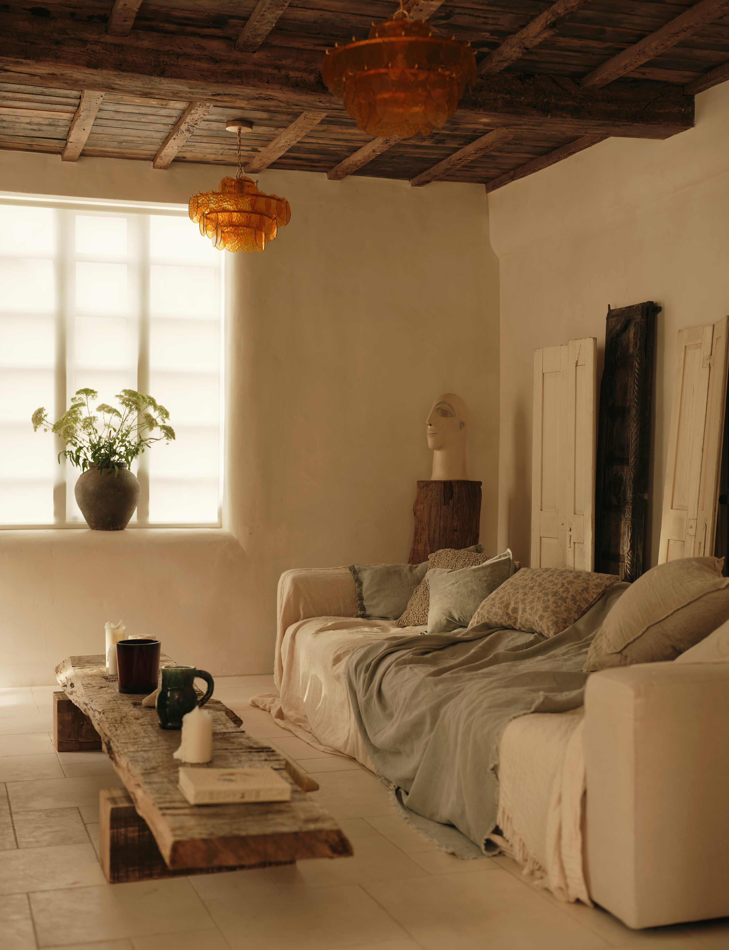 A living room with a white sofa, a throw blanket, and cushions on top. There is a large, long coffee table in the middle with mugs and candles on top. On the windowsill, in a plant inside a vase