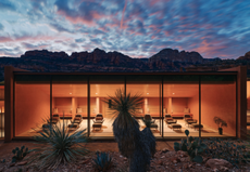 The image shows a relaxation room at Mii Amo spa, lit up at sunset. In the foreground are cacti and in the background, mountains and a multi-colored sunset.