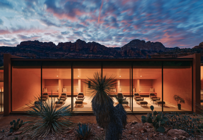 The image shows a relaxation room at Mii Amo spa, lit up at sunset. In the foreground are cacti and in the background, mountains and a multi-colored sunset.