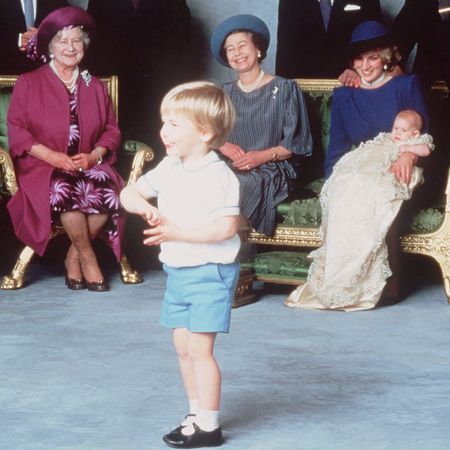 A toddler Prince William wearing a white shirt and blue shorts standing in front of Queen Elizabeth, Princess Diana and the Queen Mother laughing as they pose in chairs