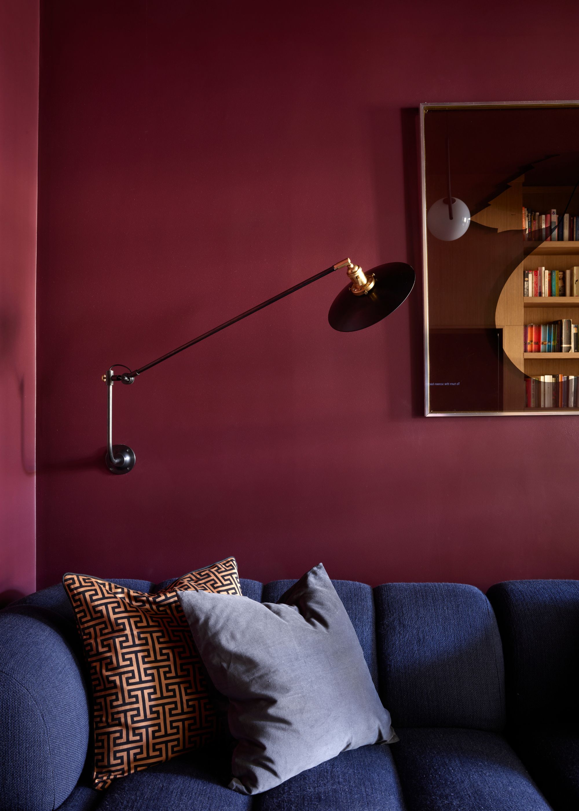 A living room painted maroon red with a frame hanging on a wall. There is also a blue colored sofa with cushions on top