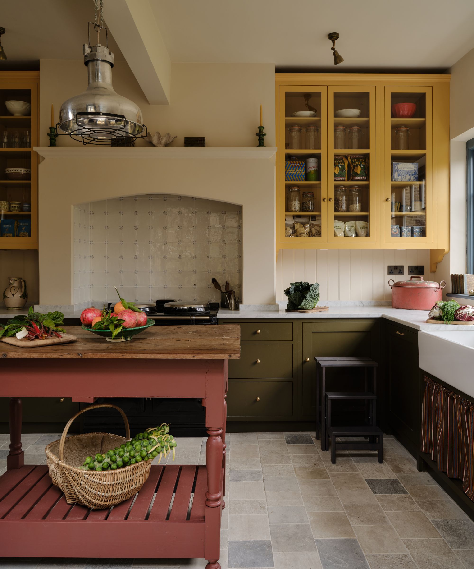 Farmhouse kitchen with dark olive low cabinets, a farmhouse sink, upper glass cabinets painted butter yellow, a large hearth with vintage cream tiles, and a red freestanding island.