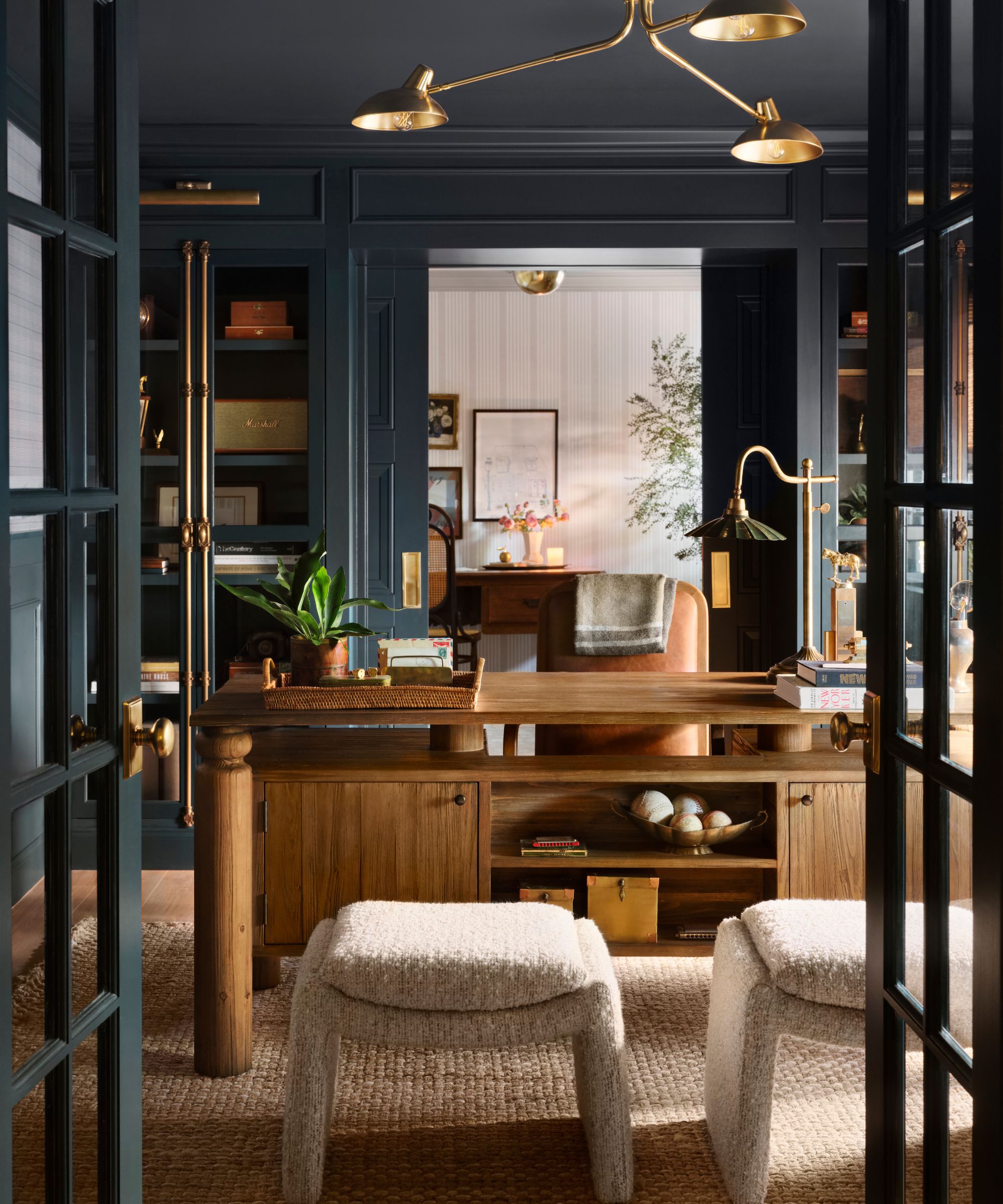 Home office with navy painted walls, wood desk, gold pendant light, leather chair and boucle stools