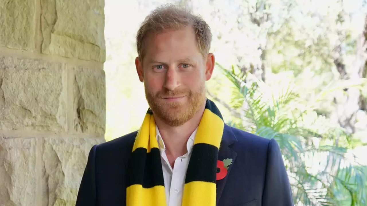 Prince Harry wearing a black and yellow scarf and a blue blazer with a poppy pin standing in front of trees 