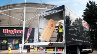 An employee unloading a Hoover fridge from the back of a Hughes delivery truck parked in front of a Hughes store