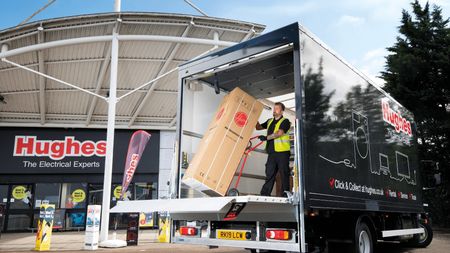 A Hughes employee unloading a hoover from the back of a Hughes delivery truck which is parked in front of a Hughes store