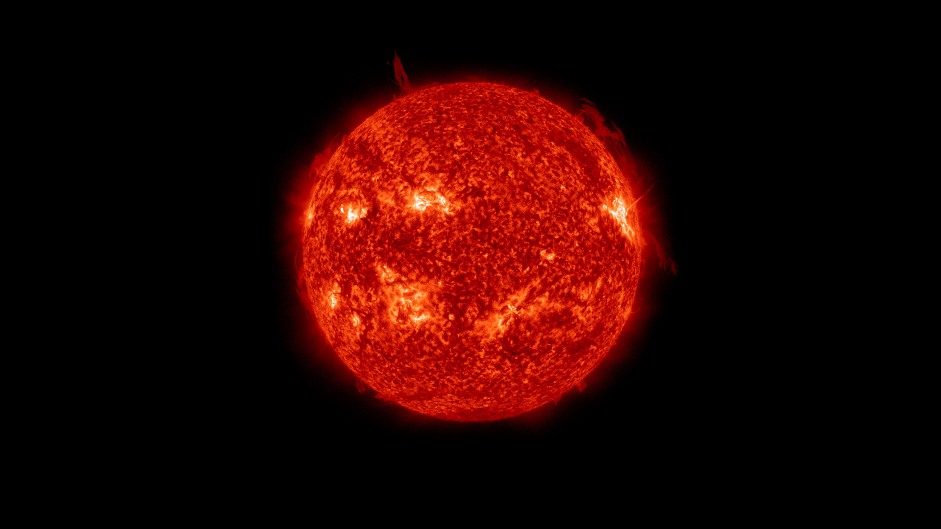 An image of the sun in space. It appears as a deep red/orange sphere with bright regions and some wispy filaments reaching out into space.