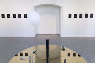 round glass mirror surface with object placed on it , in a room with white walls and green floor. Black Portrait posters on the white wall.