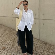 Débora Rosa wearing a white shirt, black trousers, flip-flops, sunglasses and a yellow shoulder bag.