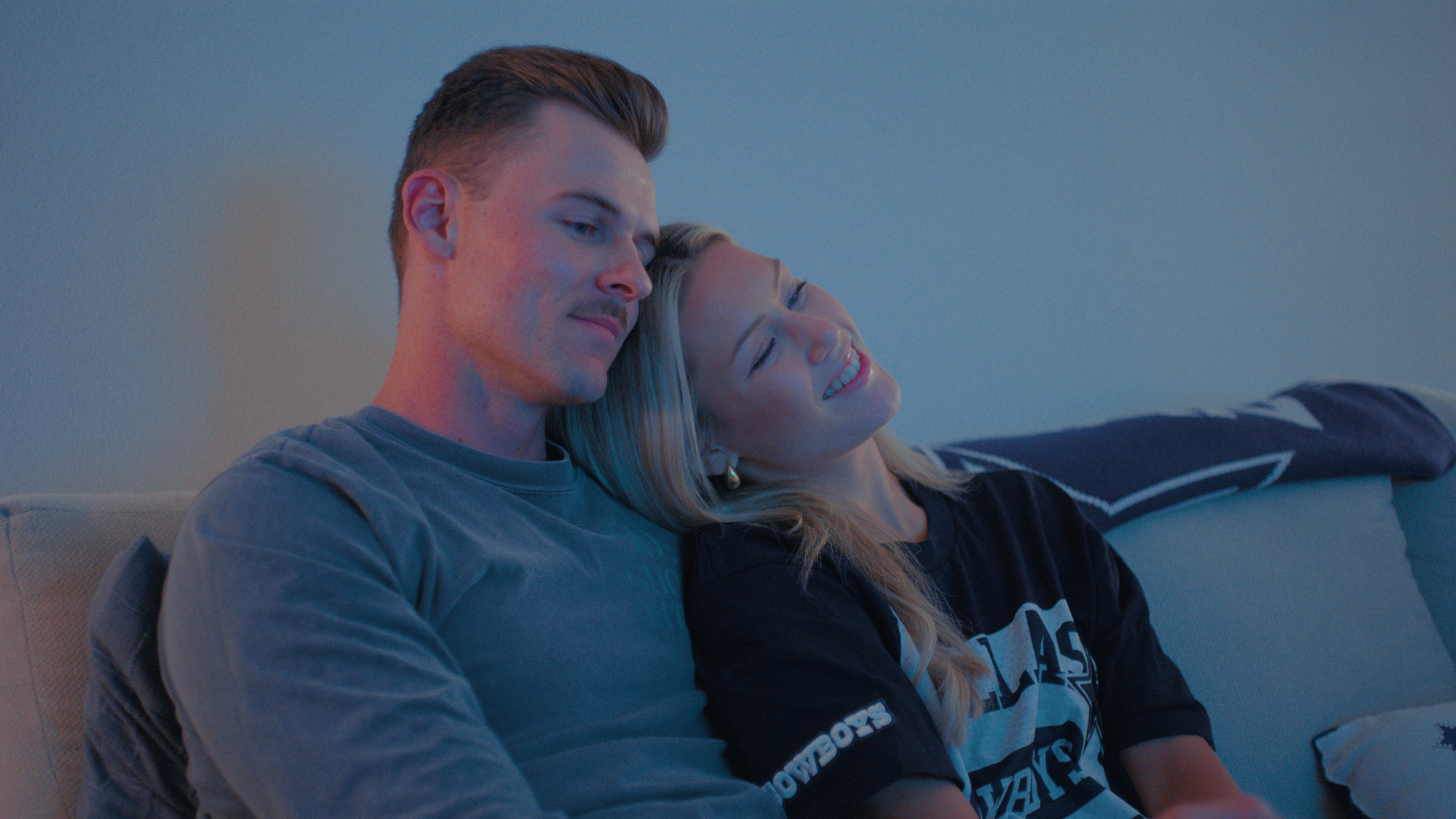 alex and megan leaning on each other sitting on a couch in america's sweethearts dcc season 2