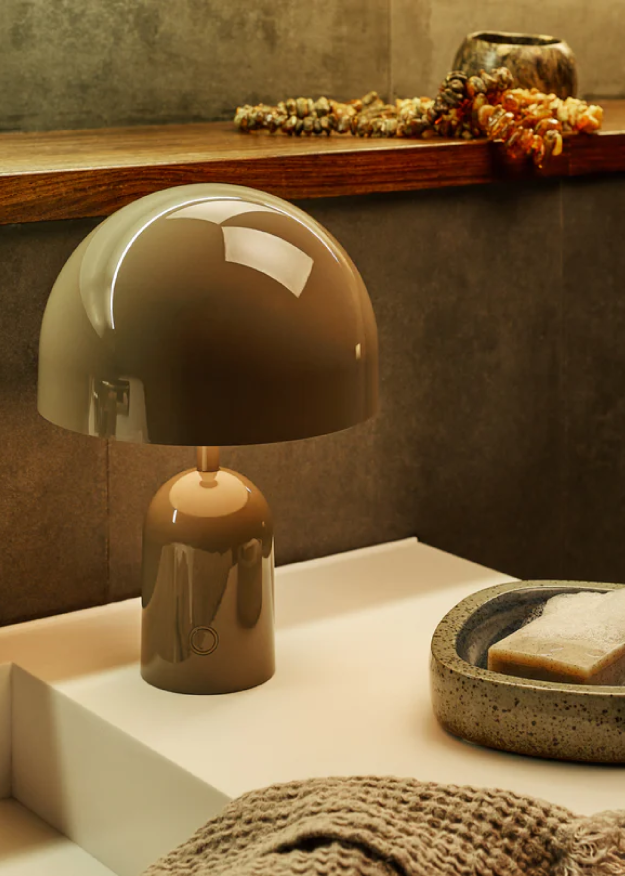 A khaki colored portable lamp on a sink basin next to a marble soap tray and towel