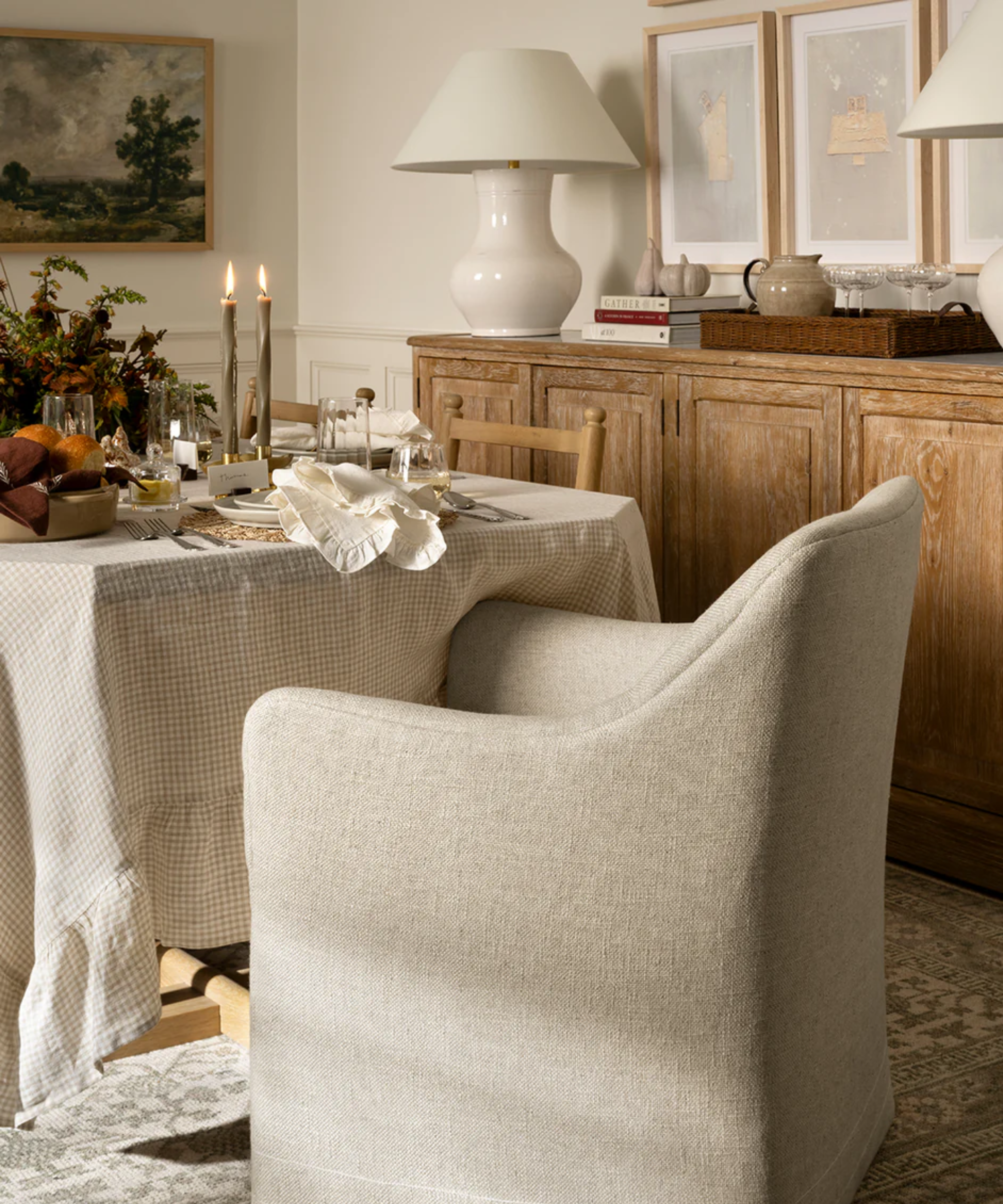 a dining room scene with a dining chair, a table covered with a gingham tablecloth, plates, utensils, napkins, glassware, and candles, and a credenza with a large lamp, books, a pitcher, and more glassware