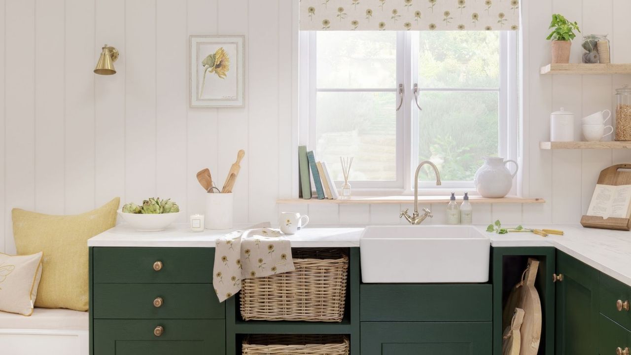 A white farmhouse kitchen sink beneath a window with green cabinets and bee patterned tea towel