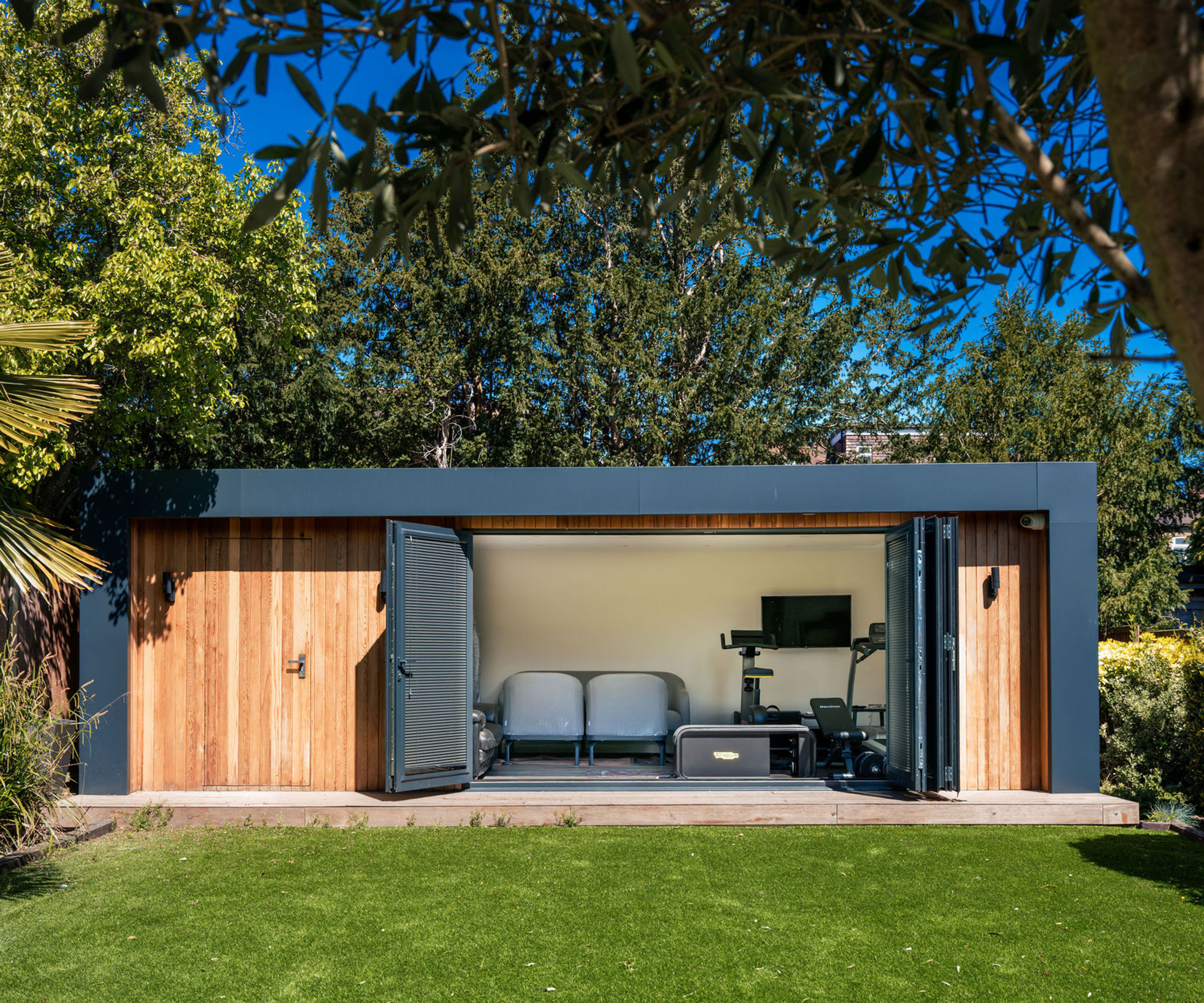 modern garden room building at the end of a garden fitted out as a home gym space. Features bifolding doors that are fully open