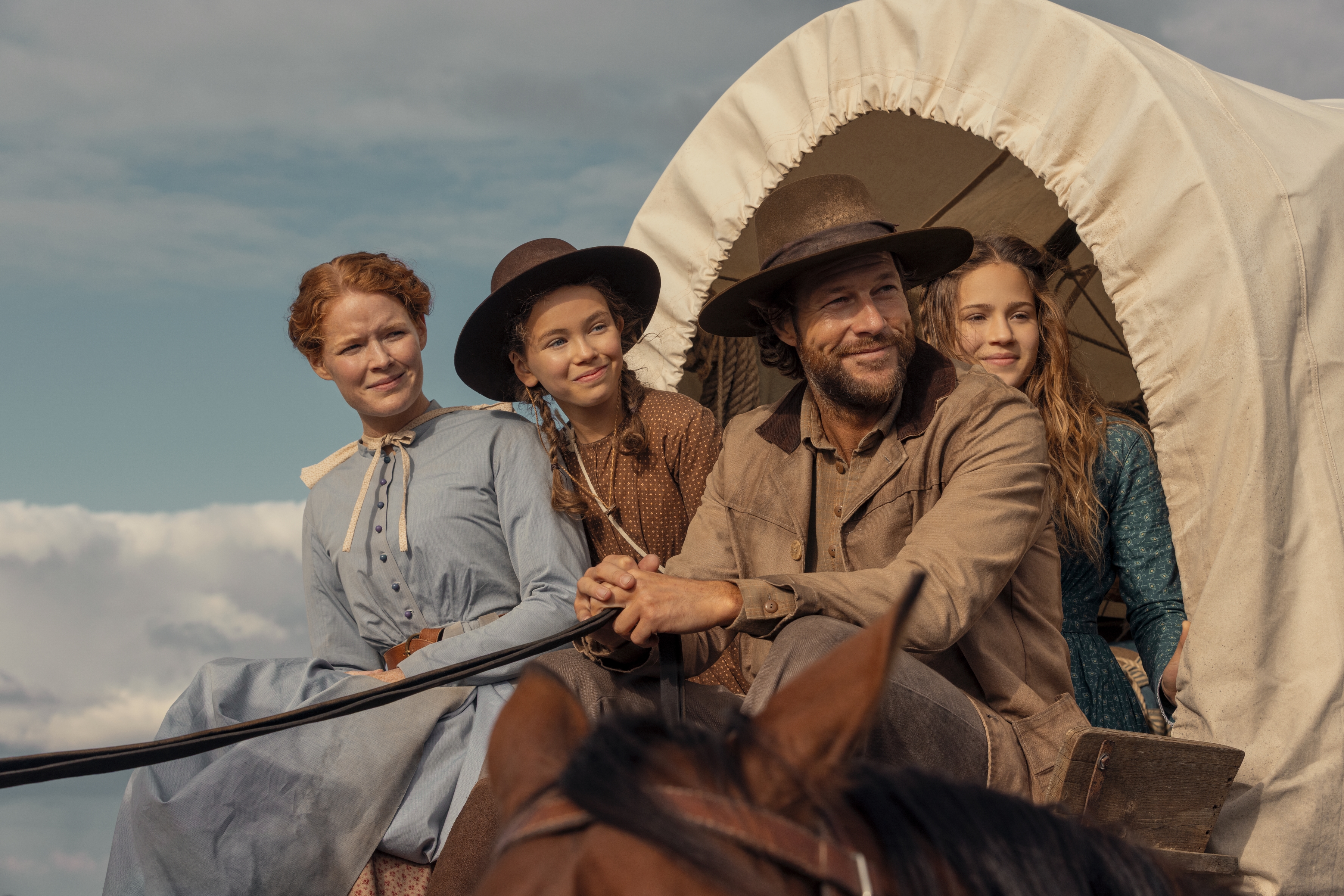 Crosby Fitzgerald as Caroline Ingalls, Alice Halsey as Laura Ingalls, Luke Bracey as Charles Ingalls, Skywalker Hughes as Mary Ingalls smile while sitting on a covered wagon in 'Little House on the Prairie.'