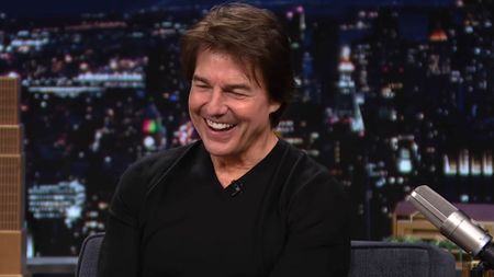 Tom Cruise is shown laughing on The Tonight Show with Jimmy Fallon, wearing a black shirt with the cityscape backdrop seen in the background.