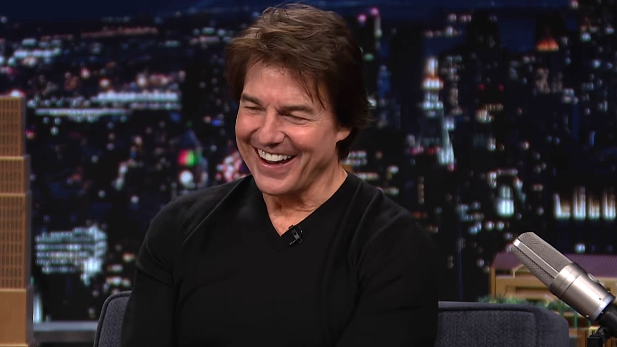 Tom Cruise is shown laughing on The Tonight Show with Jimmy Fallon, wearing a black shirt with the cityscape backdrop seen in the background.