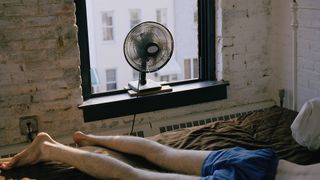 Man lying face down on a bed with fan pointed in his direction