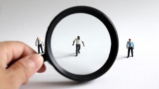 A hand holds a magnifying glass up against a model of a black employee, while two white employees flank outside the view of the lens against a white background
