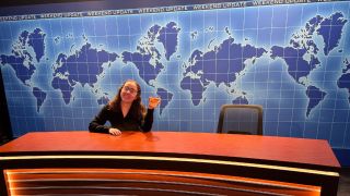 Riley Utley sitting in the left Weekend Update chair behind the desk, she's smiling and holding a glass in her right hand.