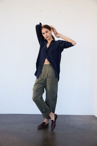 a model wears a black shirt and olive green pants with Jamie Haller shoes in front of a plain wall