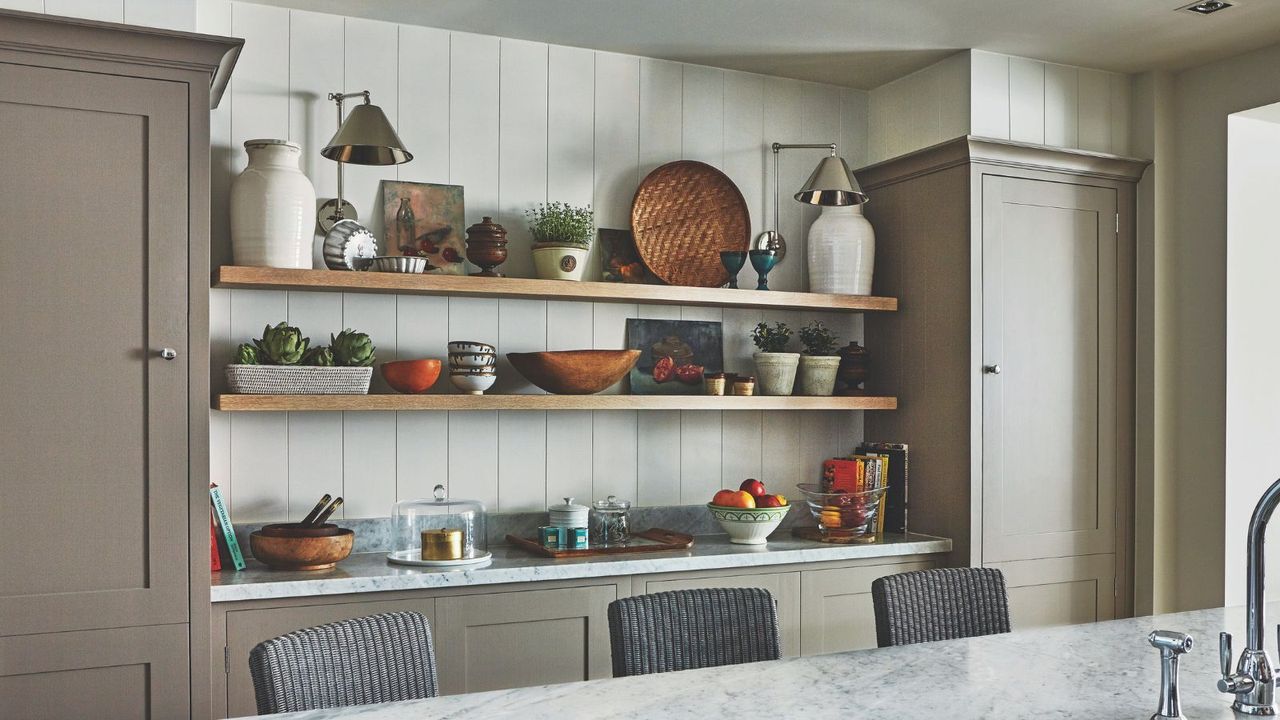 Wooden shelves in a kitchen filled with eclectic plants, bowls, vases, lamps and glassware. On either side are brown cupboards, and in the foreground is a marble island with grey rattan chairs, and a silver faucet is seen to the bottom right hand corner.