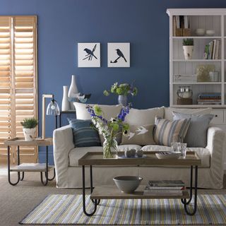 living room with blue walls, plantation shutters, white couch with blue striped cushions and industrial look coffee and side tables