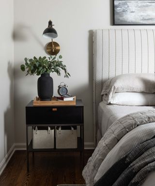 corner of bedroom with white walls and striped white headboard, gray bedding and black nightstand with vase of green foliage