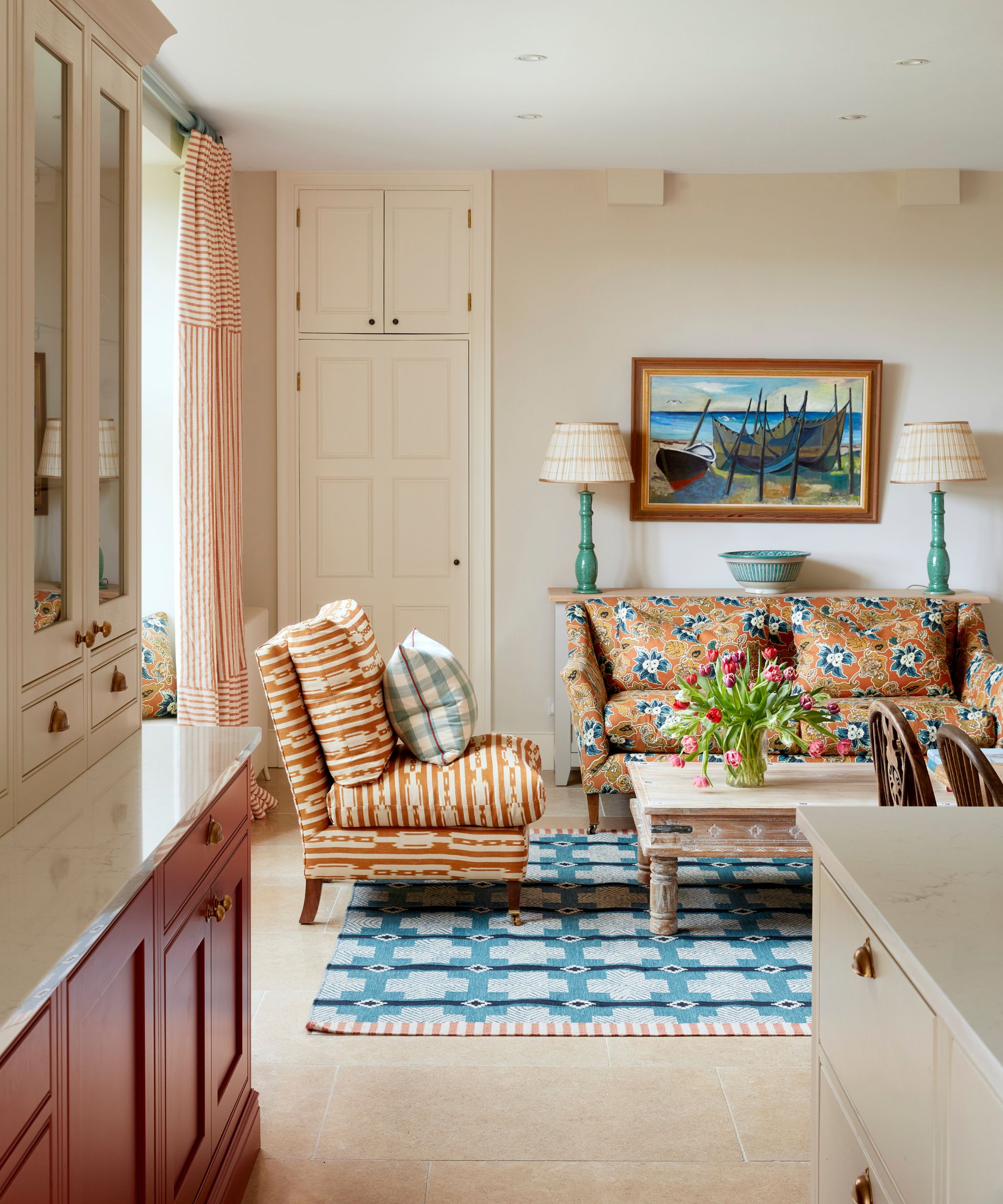 a soft neutral snug area in an open plan soft kitchen with a blue flatweave rug, orange floral sofa, orange ikat print chair and rustic coffee table