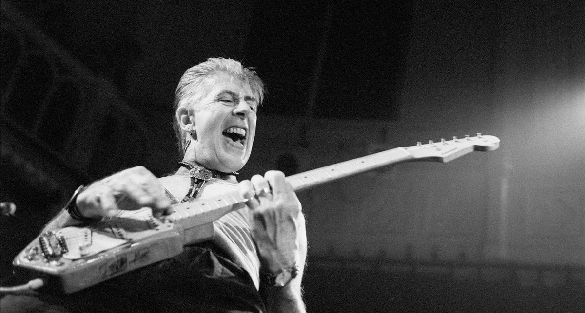 A black and white action shot of John Mayall onstage playing electric guitar in 1991