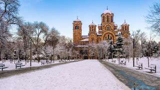 A snow-covered park with benches leads to an ornate church, surrounded by trees glistening with frost against a clear blue sky