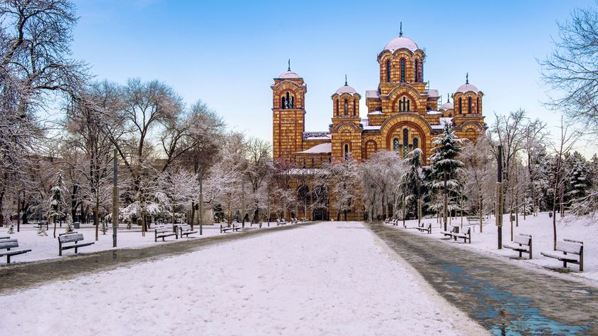A snow-covered park with benches leads to an ornate church, surrounded by trees glistening with frost against a clear blue sky