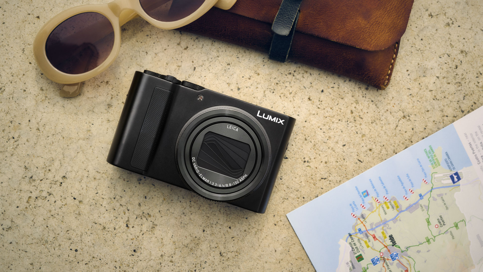 Panasonic Lumix TZ300 travel compact camera on a stone surface alongside a map and travel bag and sunglasses