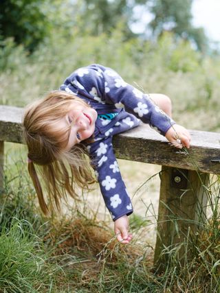 A photo of a child lying over a bench with wild grass around her.