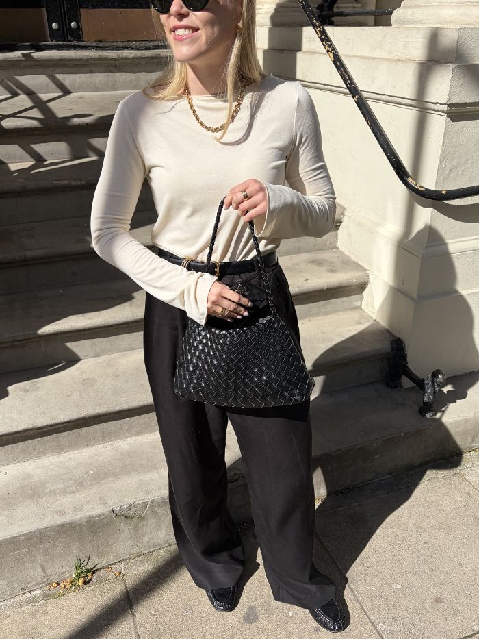 Woman wears white top, black tailored trousers, loafers and carries black woven bag