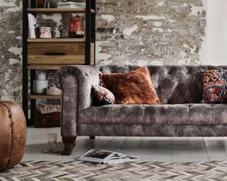 A grey velvet sofa in a living room with stripped brick walls and bookcase