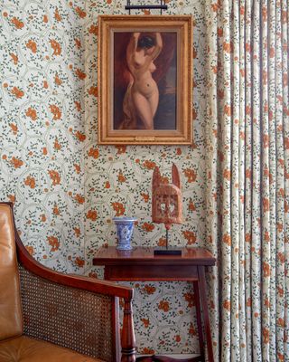 Matching wallpaper and curtains in ditsy floral pattern, antique artwork, ornaments on wooden side table, wood, leather and rattan chair