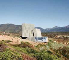 Ocho Quebradas Concrete House, glass fronted, a person stood looking out of the floor to ceiling windows, building set in a hilltop, surrounding landscape of moss, shrubs and mountainous backdrop, blue daytime sky 