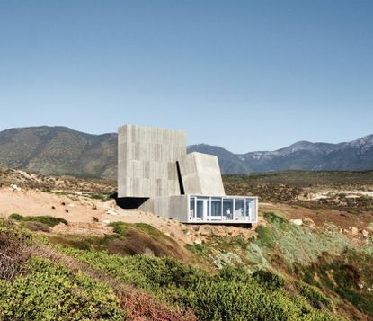 Ocho Quebradas Concrete House, glass fronted, a person stood looking out of the floor to ceiling windows, building set in a hilltop, surrounding landscape of moss, shrubs and mountainous backdrop, blue daytime sky 