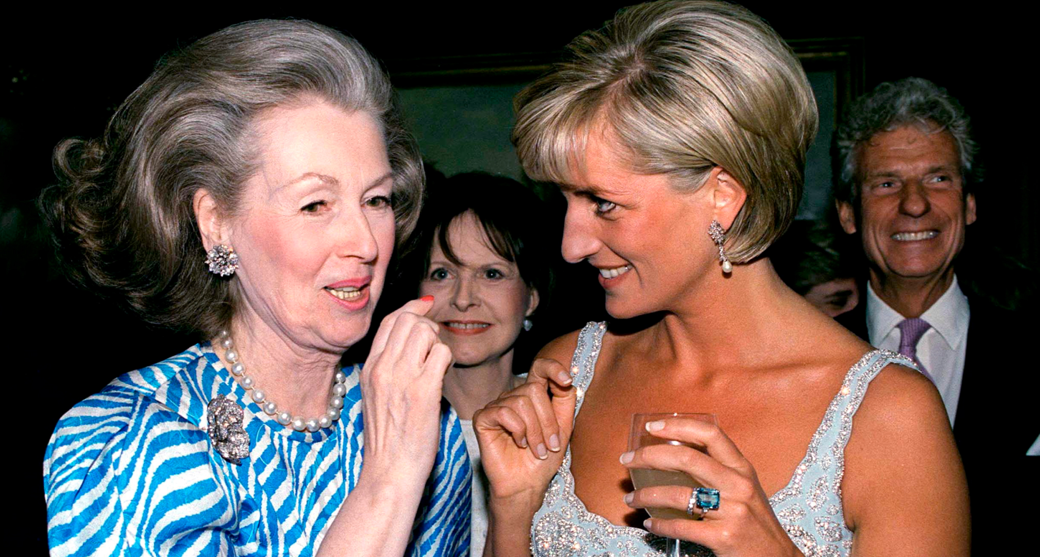 Princess Diana wearing a blue dress, holding a wine glass and smiling at her stepmother, Raine Spencer