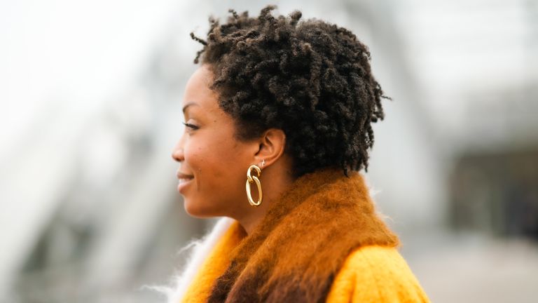 Ellie Delphine wears golden earrings, a multi colored wool Loewe scarf, an orange wool coat with from Mariam Al Sibai, on January 06, 2021 in Paris, France.