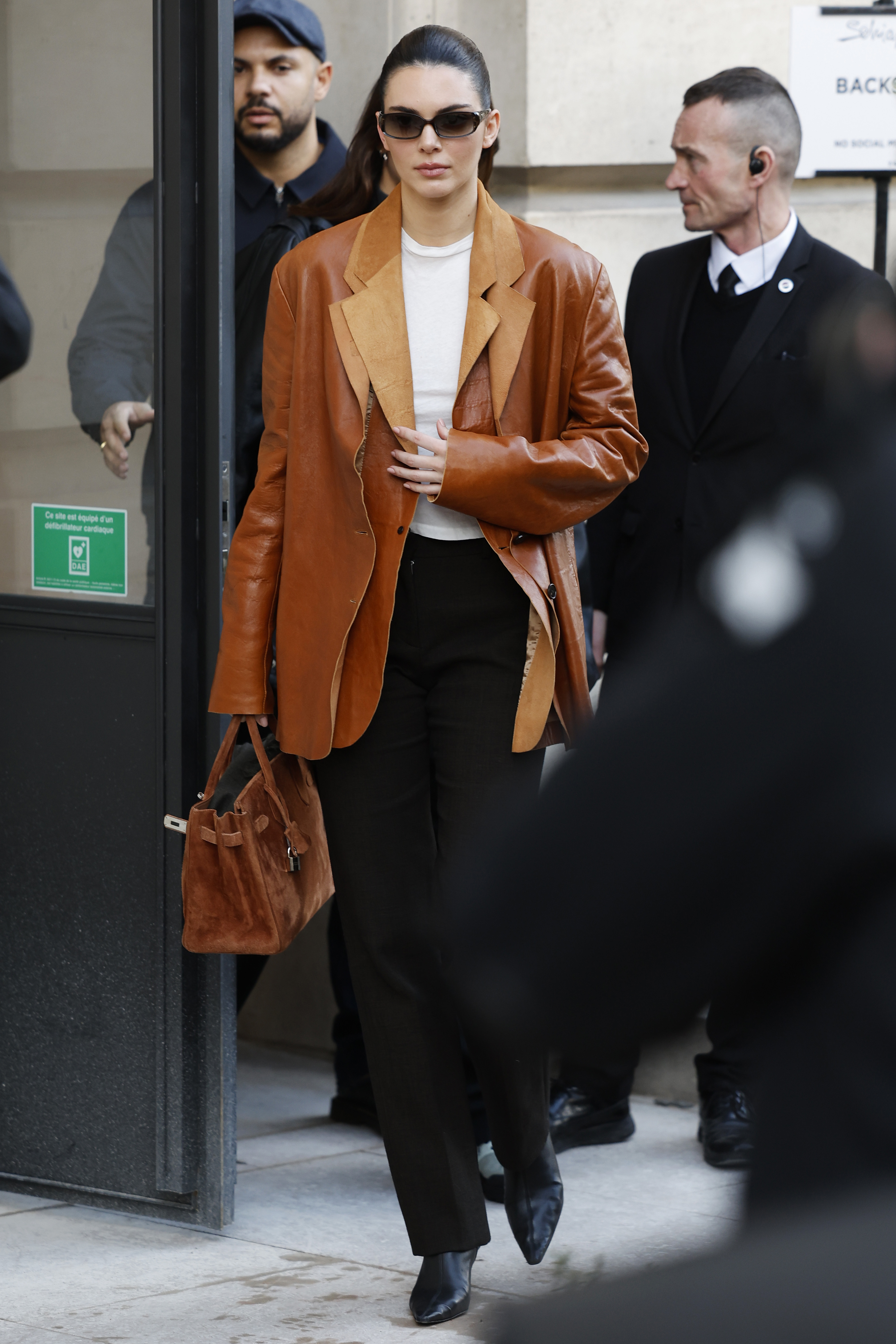 Kendall Jenner wears a white top, brown leather double blazer, brown suede Hermès bag, black trousers, black boots, and black sunglasses outside Schiaparelli during Haute Couture Spring-Summer 2025 as part of Paris Fashion Week on January 27, 2025 in Paris, France.