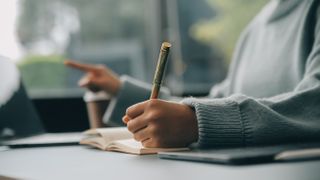 Woman sitting at a table, and working on a laptop and writing in a notebook