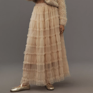 A close up of a model wearing a cream tulle skirt with gold ballet shoes shot against a dark background