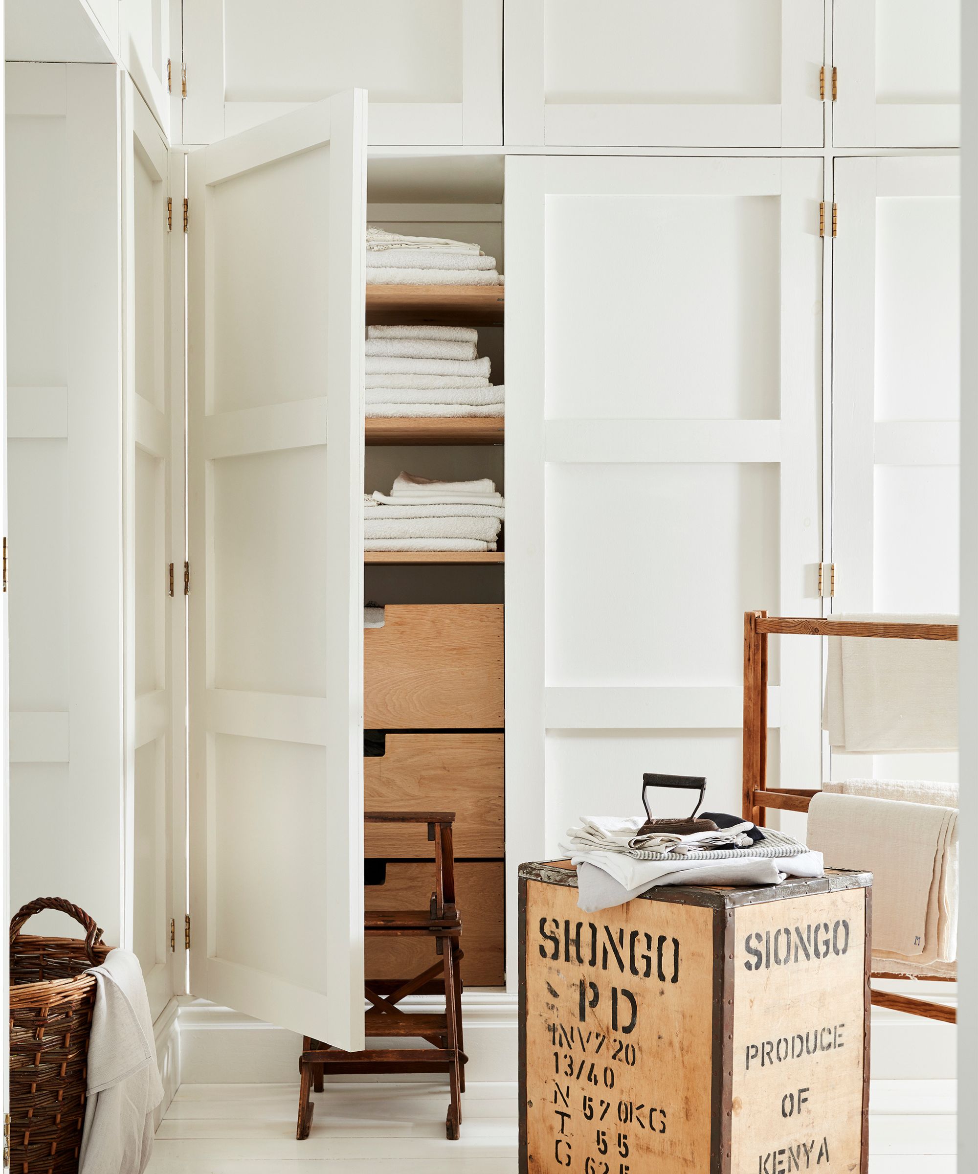 White cupboards built-in to walls, one door opening revealing a linen cupboard stacked with towels and bedding, a wooden footstool and towel rack beside it