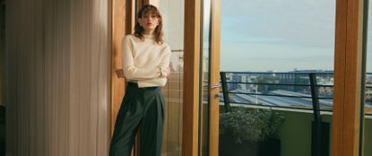 Model wearing a white mock neck long sleeve and green tailored trousers by open balcony doors of an apartment